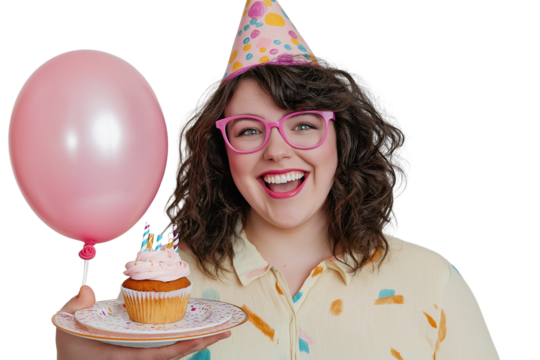 Portrait of a smiling young woman wearing pink glasses and a party hat, holding a balloon. She is also holding a plate with a cupcake and a candle on a transparent background