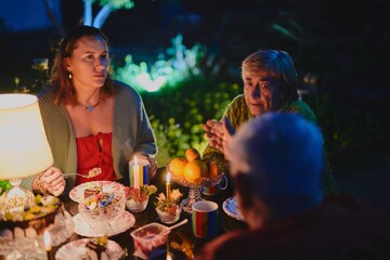 Happy elderly couple and their adult daughter celebrate holiday in the courtyard of the house, family of three have dinner in lamplight