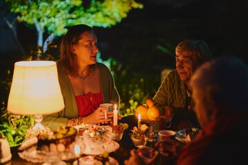 Happy elderly couple and their adult daughter celebrate holiday in the courtyard of the house, family of three have dinner in lamplight