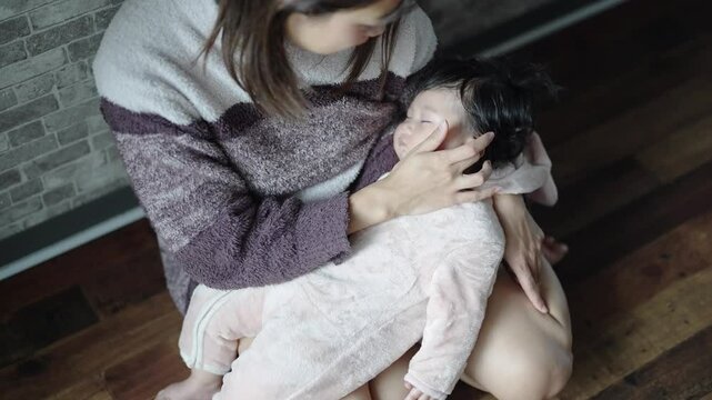 In a warm winter Japanese apartment room, a 6-month-old Japanese girl wearing fluffy clothes and her mother are sitting on the floor holding their sleeping baby. Slow motion video.
