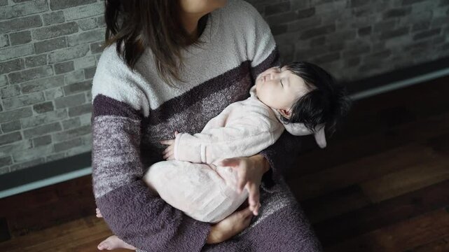 In a warm winter Japanese apartment room, a 6-month-old Japanese girl wearing fluffy clothes and her mother are sitting on the floor holding their sleeping baby. Slow motion video.