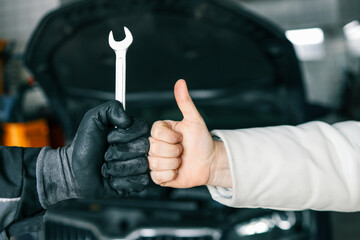 Close-up of a mechanic's gloved hand holding a wrench and a customer's hand giving a thumbs-up, symbolizing satisfaction and trust in car repair services