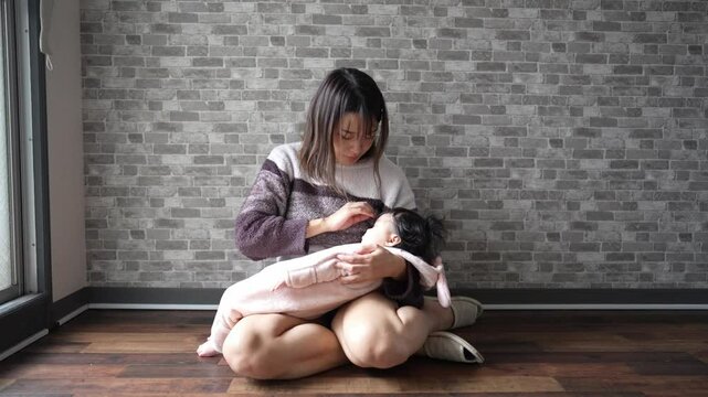 In a warm winter Japanese apartment room, a 6-month-old Japanese girl wearing fluffy clothes and her mother are sitting on the floor holding their sleeping baby. Slow motion video.