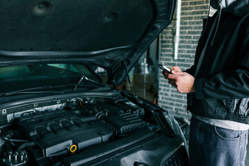 Mechanic using a smartphone while inspecting an engine under the open hood of a car in a professional garage. Modern technology in automotive diagnostics and repair