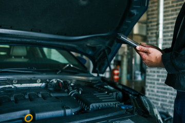 A mechanic holding a tablet while inspecting a car engine in a workshop. The focus is on the...