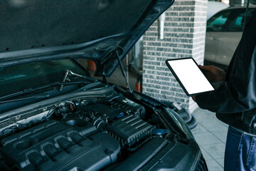 A tablet with a blank screen held by a mechanic above a car engine, symbolizing modern diagnostic tools for vehicle maintenance and repair. The engine is visible in the background