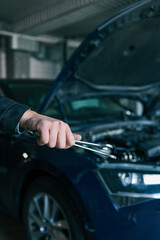 A mechanic holding wrenches in front of an open car hood in a garage. The image emphasizes automotive repair tools and professional car maintenance in a workshop setting