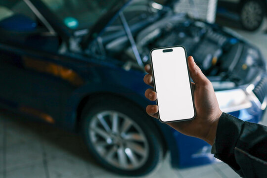 Hand holding a smartphone with a blank screen in front of an open car hood, showing the engine compartment. Concept for car diagnostics, automotive apps, or remote vehicle monitoring - Powered by Adobe