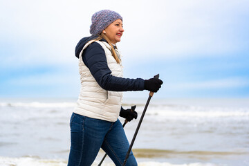 Beautiful middle aged woman training Nordic walking on sandy beach in winter. Side view	