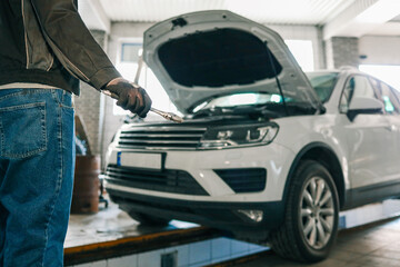 Mechanic holding an adjustable wrench in a workshop. A white car with its hood open is visible in the background, indicating repair or maintenance work