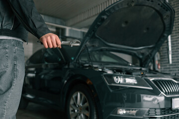 Hand holding an adjustable wrench in an automotive repair shop. A blue car with an open hood is visible in the background, highlighting repair or maintenance activity