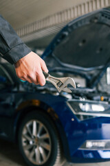Hand holding an adjustable wrench in an automotive repair shop. A blue car with an open hood is...