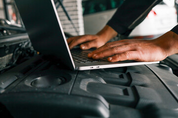 Close-up of a mechanic's hands typing on a laptop placed on a car engine in a workshop, showcasing the integration of technology in vehicle diagnostics and repairs