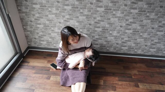 In a warm winter Japanese apartment room, a 6-month-old Japanese girl wearing fluffy clothes and her mother are sitting on the floor holding their sleeping baby. Slow motion video.