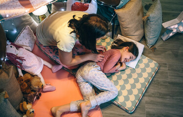 Woman waking up a smiling young girl sleeping lying over cozy mats at home. Mother and happy...