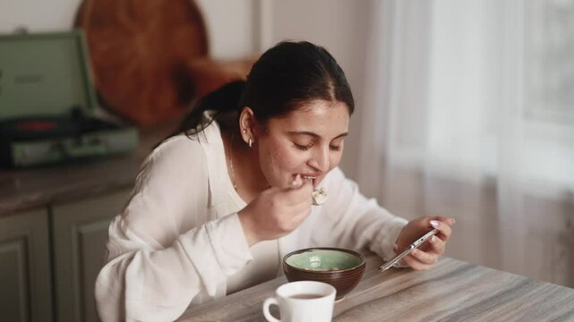 Smiling young indian woman enjoying beautiful morning with tasty breakfast hold smartphone watching online videos entertainment program in social media alone in kitchen at home