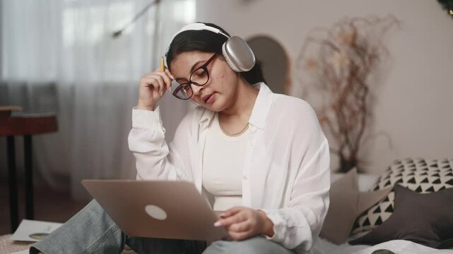 Boring unmotivated young indian woman student listening online lecture by laptop computer sitting on bed at home Tired girl with uninteresting lesson on remote distance education indoors