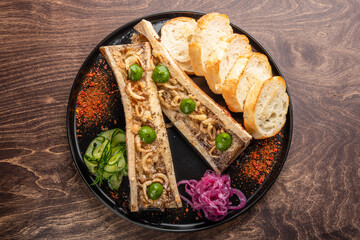 A delicious serving of bone marrow topped with herbs, accompanied by toasted bread, pickled onions, and sliced cucumbers, presented on a black plate against a wooden background.