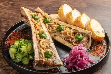 A delicious serving of bone marrow topped with herbs, accompanied by toasted bread, pickled onions, and sliced cucumbers, presented on a black plate against a wooden background.