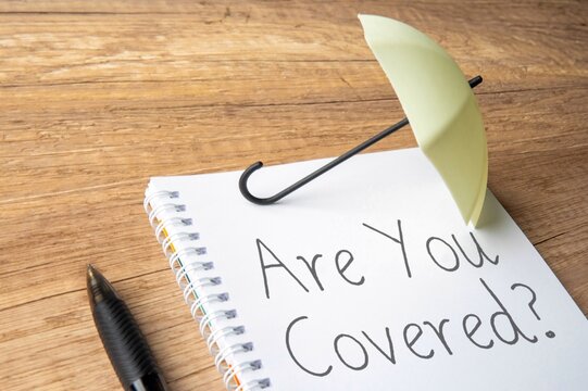 A small umbrella is placed on top of a notebook with the words "Are You Covered?". Protection, insurance, financial security concept.