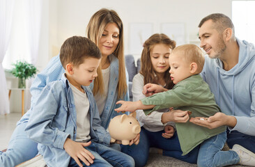Happy large family teaching children about saving money, placing coins into piggy bank while...