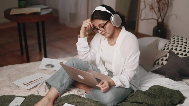 Boring unmotivated young indian woman student listening online lecture by laptop computer sitting on bed at home Tired girl with uninteresting lesson on remote distance education indoors
