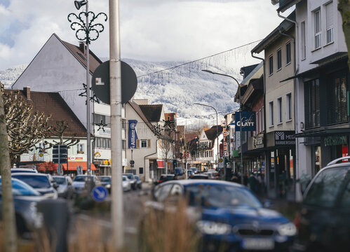 Achern, Germany - Jan 3, 2025: A bustling town street with shops, vehicles, and people, surrounded by snow-covered hills under a cloudy winter sky in Achern, germany