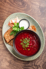 A traditional bowl of borscht served with sour cream, fresh herbs, dark bread, and cured meat on a decorative plate, set on a wooden table. A vibrant and hearty meal.
