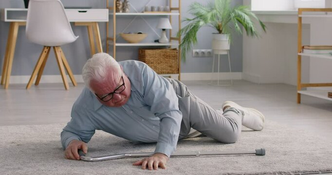 Senior elderly man falling down and lying on the floor with walking stick in living room at home alone and trying to get up with the help of his crutches suffering with pain. 4k video.