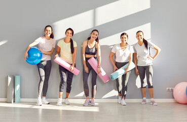 Fototapeta premium A group of athletic girls with yoga mats stand against the wall in the gym, portrait. Happy sportswomen smiling. Joy and satisfaction from yoga. Concept of health and perseverance in sports