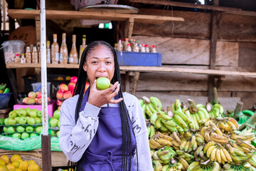 A young black african woman smiling while eating an apple in an outdoor fruit market with various...