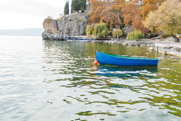Tranquil scene of a small blue boat moored on the calm waters of a lake, with a picturesque landscape of autumn colorful trees in the background.