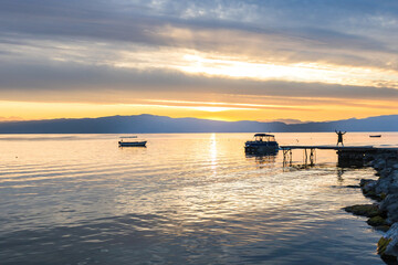 Breathtaking sunset over Lake Ohrid, with pier and a small boat anchored in the calm waters. The...