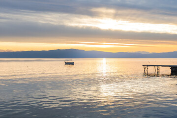Fototapeta premium Breathtaking sunset over Lake Ohrid, with pier and a small boat anchored in the calm waters. The golden light reflecting on the water creates a serene and picturesque scene.