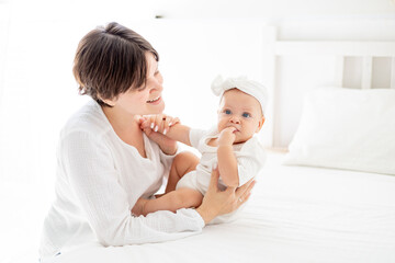 mom gently hugs her baby daughter kissing and admiring them, happy family, mom with baby on the bed, maternal love and care, mom on maternity leave playing with the baby