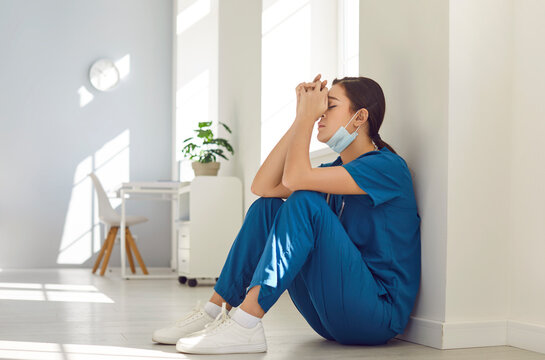 Portrait of tired depressed female nurse wearing blue face mask and uniform sitting on hospital floor in hallway. Young exhausted woman doctor stressed from hard work in medical clinic.
