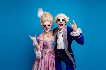 A playful couple in baroque costumes and sunglasses pose against a vibrant blue backdrop, blending...
