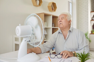 Senior man suffering from summer heat and trying to survive one more hot day with electric fan....