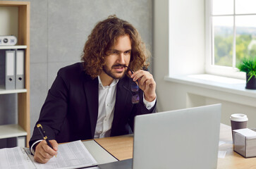 Obraz premium Portrait of young attractive business man with long hair working on a laptop at office or at home, looking on a computer screen and writing, considering new projects or startups, analyzing company.