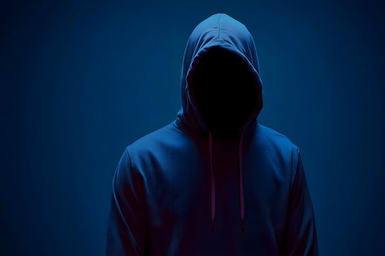 Hacker man is wearing a hoodie and is standing in front of a dark blue background with copy space. Concept of mystery and anonymity, as the man's face is hidden behind the hood