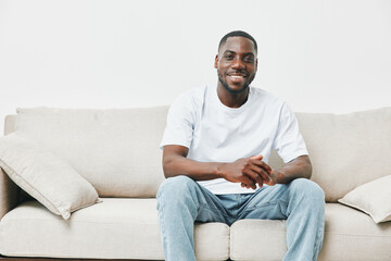 Happy young African American man sitting on a light beige sofa, wearing a casual white t shirt and jeans, conveying a relaxed vibe and confidence