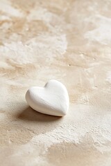 A delicate handmade clay heart in soft white rests on a matte beige surface, casting gentle shadows. The minimalist backdrop offers clean space ideal for text or design elements