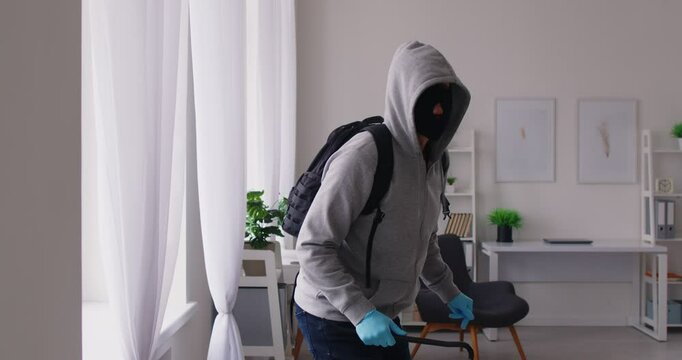 Masked thief wearing black balaclava stealthily climbing through window into apartment, committing burglary. Criminal intruder breaking into home, attempting theft and sneaking. Crime, robbery concept