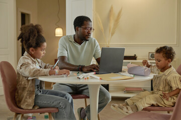 Medium shot of adult African American man working on laptop with son and daughter playing next to dad while sitting at table in cozy living room, concept of work life balance