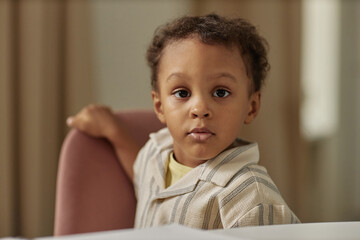 Portrait of little cute Black boy with calm expression on face looking at camera while sitting in chair at home, copy space