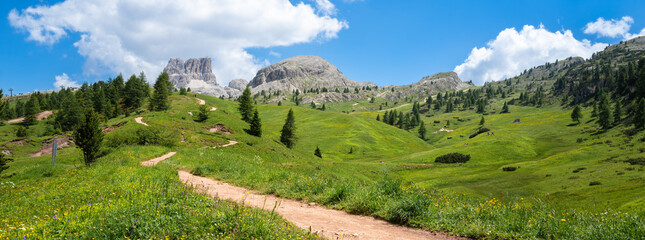 Hiking trail at Valparola mountain pass winds through lush green meadows,  views of majestic...