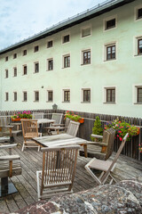 A tranquil outdoor patio scene featuring wooden furniture set against a pale green building in Kitzbuhel, Austria. Flower boxes add a touch of color. 