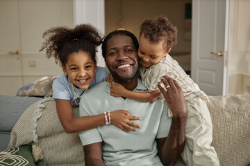 Portrait of cheerful African American father posing for camera while little kids gently hugging dad captured together on couch in living room, copy space