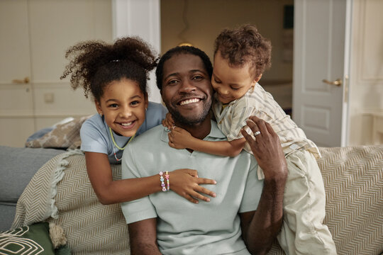 Portrait of cheerful African American father posing for camera while little kids gently hugging dad captured together on couch in living room, copy space
