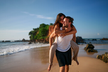 Happy couple enjoys beach getaway. Man carries woman piggyback by ocean. Playful pair laugh in sunlight, sandy shore. Casual dating, fun moments. Romantic walk, sea backdrop, holiday vibes.